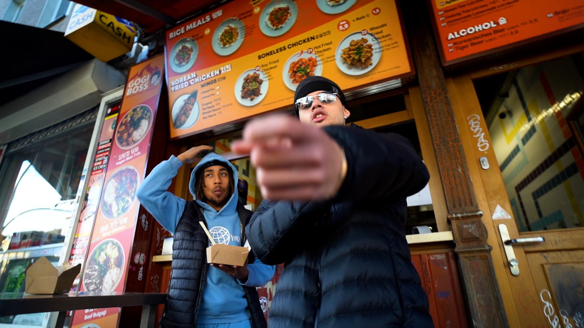 Two rappers outside a Korean takeaway during a music video shoot, one pointing into the camera