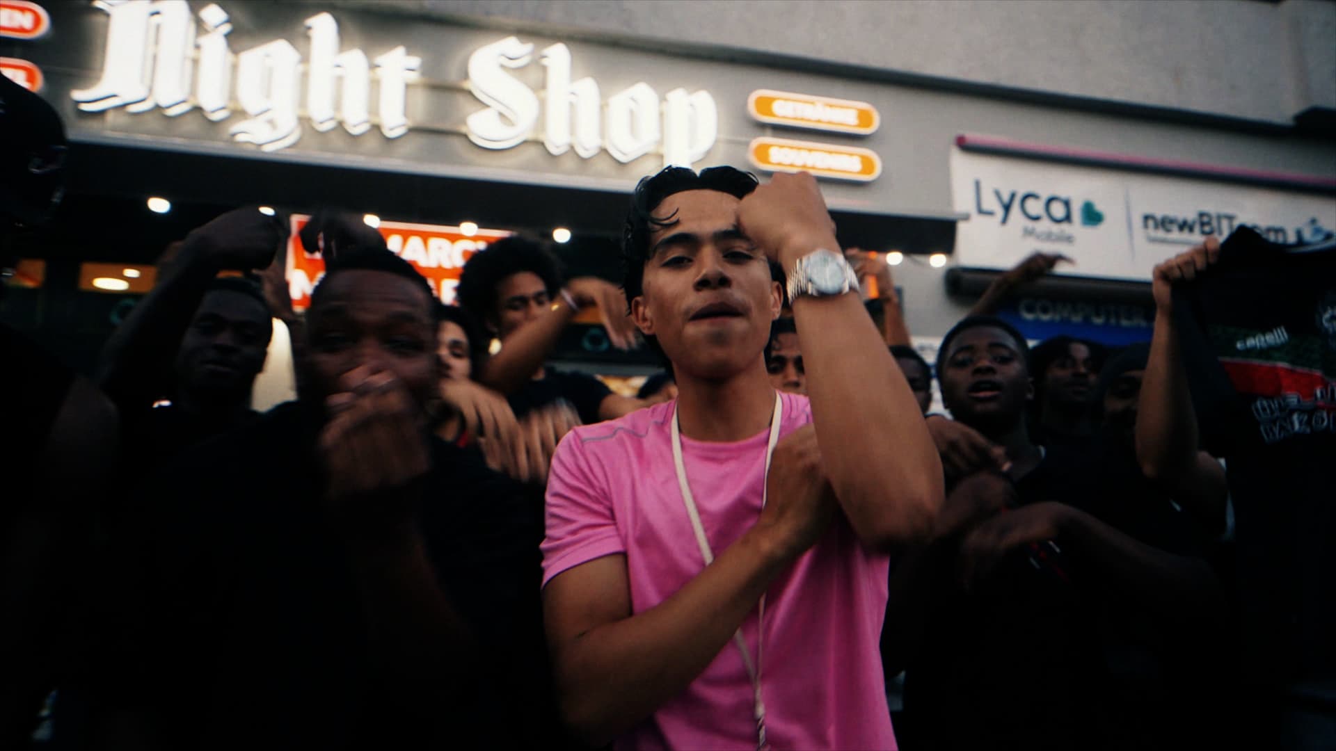 Rapper in a pink shirt performing at night in front of a lit Night Shop sign, surrounded by a crowd